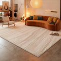 A large rectangular area rug in a light beige and off-white marble-like pattern, shown in a modern, minimalist living room with a white sofa, gray tiled floor, and a black armchair.
