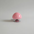 A pink mushroom-shaped table lamp with a chrome base sits on a wooden bedside table in a bedroom setting next to a bed and a framed picture. The brand name 'lamop' is printed on the lamp.