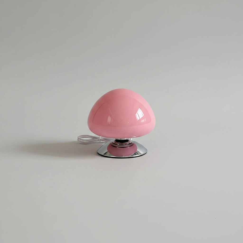 A pink mushroom-shaped table lamp with a chrome base sits on a wooden bedside table in a bedroom setting next to a bed and a framed picture. The brand name 'lamop' is printed on the lamp.