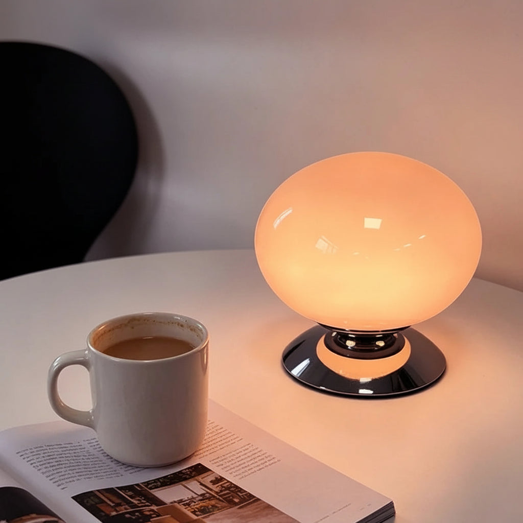 A glowing oval table lamp with a glossy black base sits on a white table next to a white ceramic mug and an open magazine. The lamp emits a warm peach-orange light.