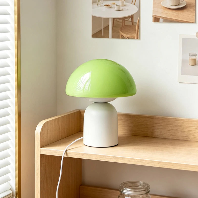 A mushroom-shaped table lamp with a glossy light green dome shade and a matte white cylindrical base, shown on a wooden shelf in a bright room.