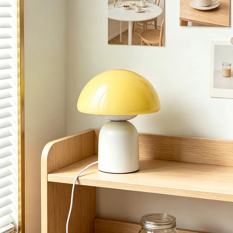 A mushroom-shaped table lamp with a glossy yellow cap and a matte white base, styled on a light wood shelf in a warm, minimalist room.