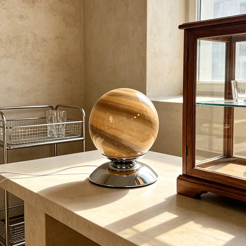 A polished spherical lamp or decorative orb with natural tan and brown banding, sitting on a reflective silver base on a light stone table in a sunlit interior.