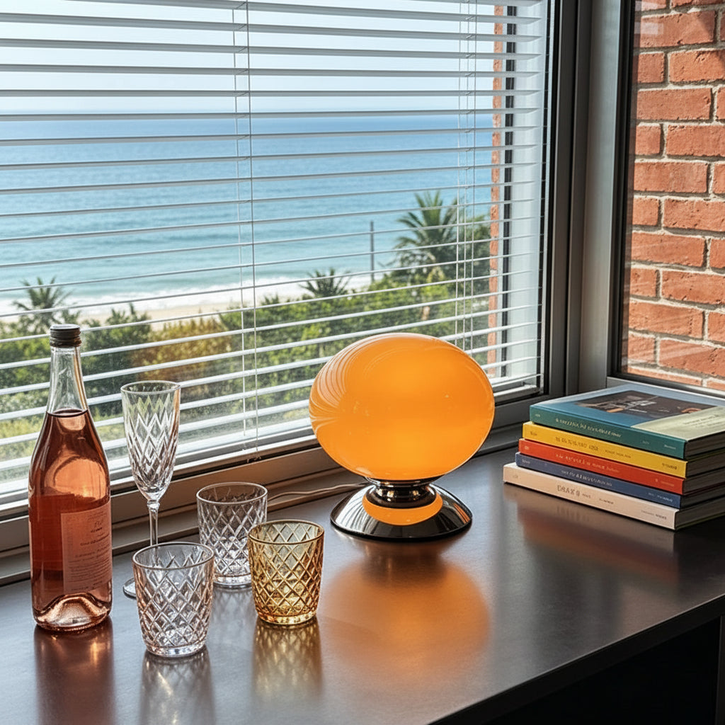 A glowing orange spherical table lamp sits on a dark windowsill beside a bottle, glasses, and a stack of books, with a beach view visible through the blinds. A circular 'Jeda Home Lighting' badge appears in the top right.