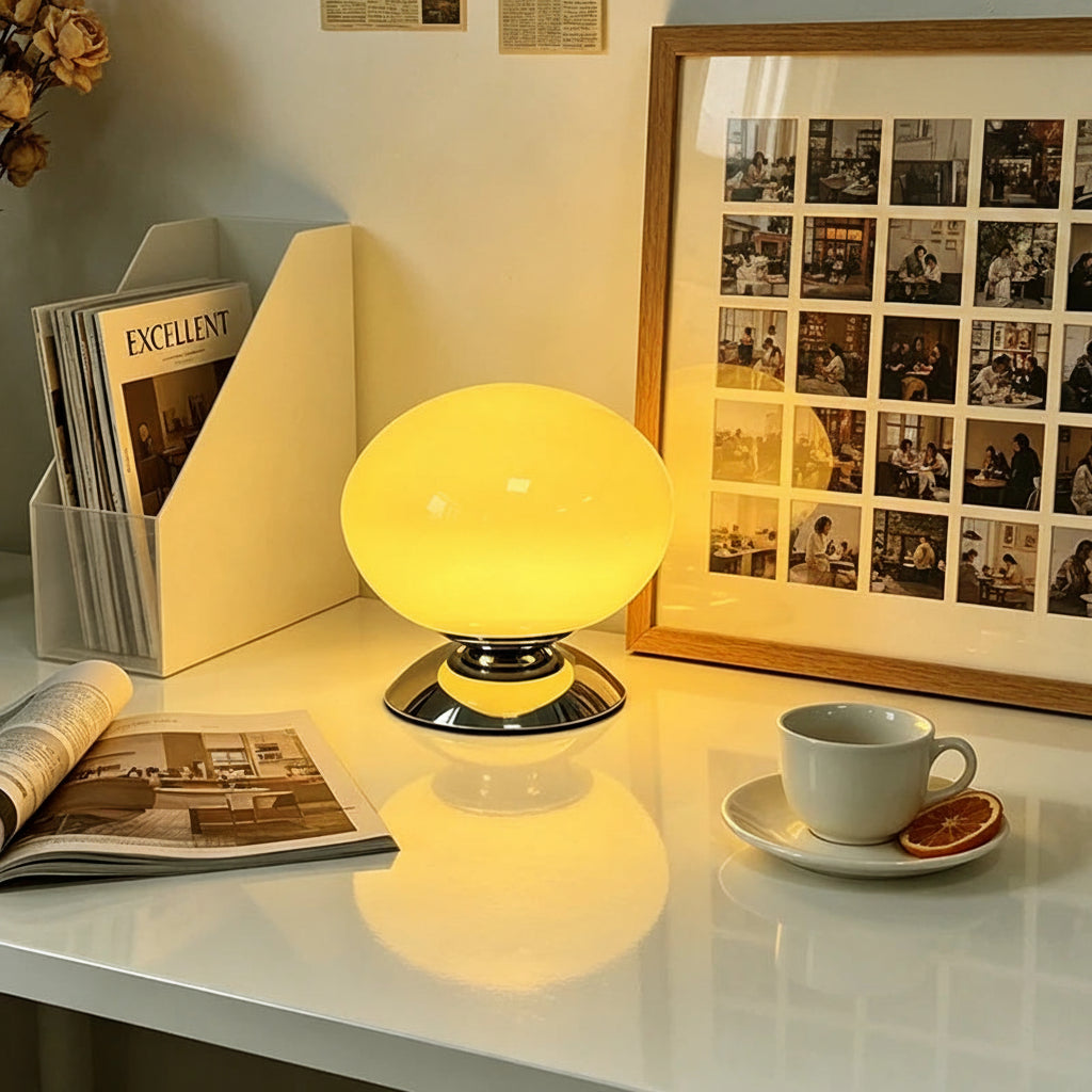 A glowing oval lamp with the brand name 'lamop' on its front, placed on a white desk next to a coffee cup, an open magazine, and a framed collage of photos. A circular badge in the top right reads 'Jieda Home Lighting'.