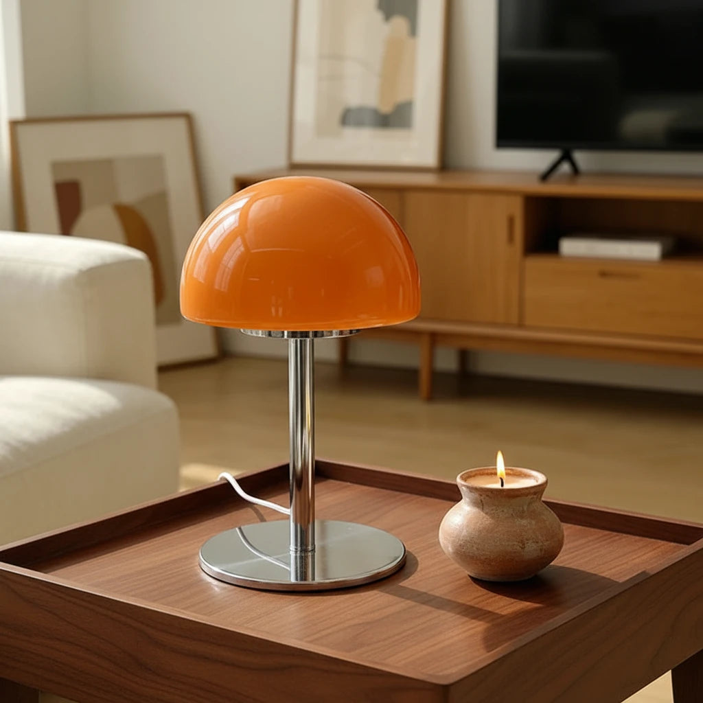 An orange mushroom-shaped table lamp with a chrome metal stem and base, shown on a round cream table in a modern living room setting next to a yellow mug.